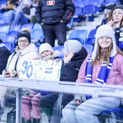 Grupa kibiców, w tym dzieci, trzymających transparent i ubranych w ciepłe nakrycia głowy, obecna na meczu piłkarskim na trybunach stadionu.