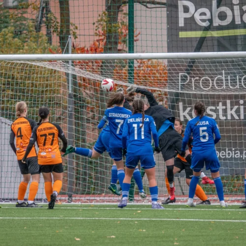 Na zdjęciu drużyna kobiet w piłce nożnej rywalizuje na boisku, a bramkarka wykonuje efektowną interwencję, odbijając piłkę na granicy bramki.
