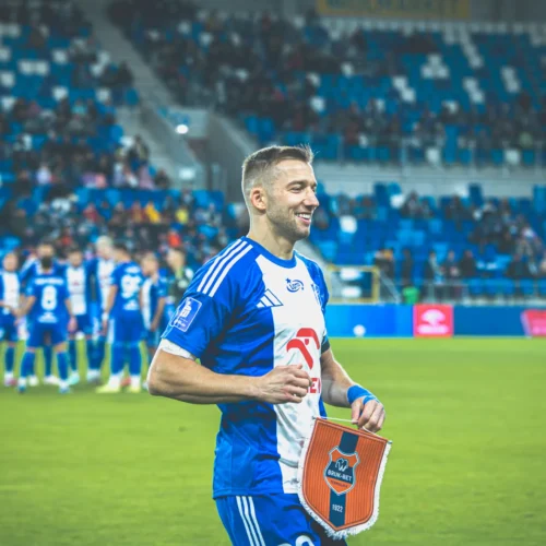 Na zdjęciu widoczny jest uśmiechnięty gracz piłkarski w niebiesko-białej koszulce, trzymający flagę drużynową, z boiskiem piłkarskim i kibicami w tle.