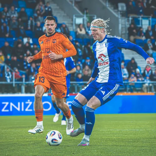 Na zdjęciu widać dwóch piłkarzy na boisku podczas meczu, jeden z nich w pomarańczowym stroju, a drugi w niebieskim, z piłką pod stopami.