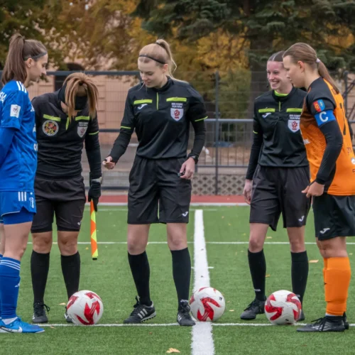 Na zdjęciu cztery kobiety w strojach piłkarskich, w tym dwie sędziny, stoją przy piłkach na boisku, przygotowując się do rozpoczęcia meczu.