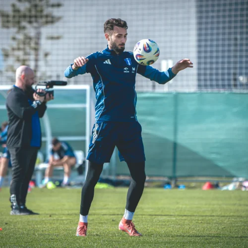 Na zdjęciu młody piłkarz trenuje na boisku, balansując piłkę na głowie, podczas gdy inna osoba nagrywa jego występ.
