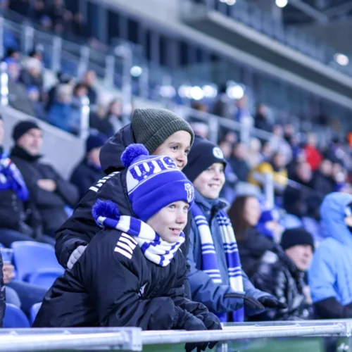 Grupa dzieci z entuzjazmem obserwuje mecz na stadionie, ubrana w cieple odzież i szaliki w kolorach drużyny.