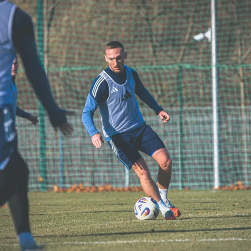 Na zdjęciu widoczny jest piłkarz trenujący na boisku, oddający się ćwiczeniom z piłką w słoneczny dzień.