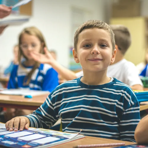 Na zdjęciu uśmiechnięty chłopiec w pasiastym swetrze siedzi przy biurku w klasie, otoczony przez kolegów i koleżanki.