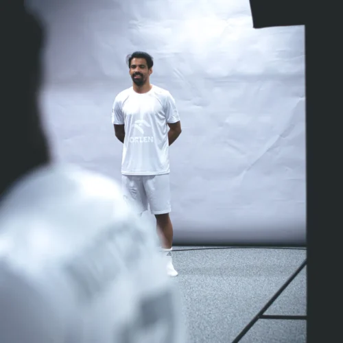 Na zdjęciu widoczny jest uśmiechający się mężczyzna w białym stroju sportowym, stojący na tle jasnej ściany w studiu fotograficznym.