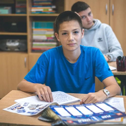 Na zdjęciu widać ucznia w niebieskiej koszulce siedzącego przy biurku w klasie, z zeszytem i przyborami szkolnymi, z kolegą w tle.
