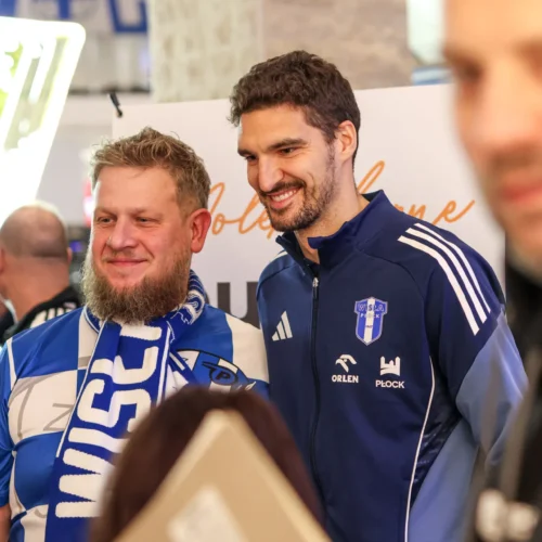 Na zdjęciu dwaj mężczyźni, jeden w niebiesko-białym szaliku, a drugi w sportowej kurtce, pozują do zdjęcia w radosnej atmosferze.