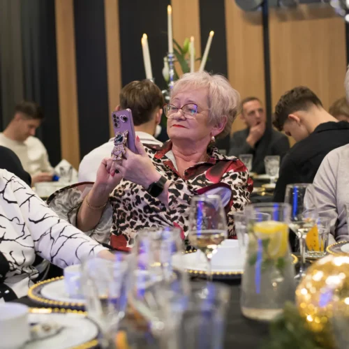 Na zdjęciu widać trzy kobiety siedzące przy elegancko zastawionym stole, z jedną z nich robiącą zdjęcie telefonem.