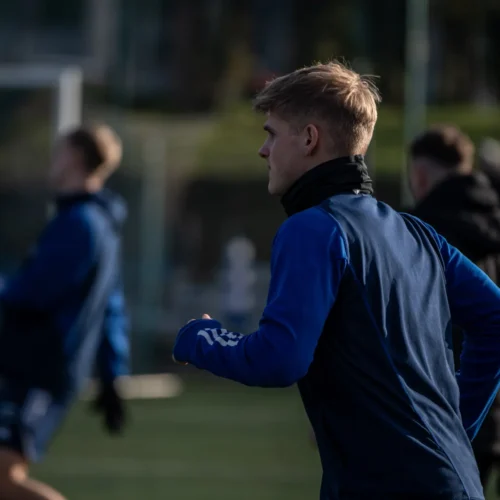 Młody sportowiec w niebieskim stroju biega na boisku treningowym, w tle widać innych zawodników.