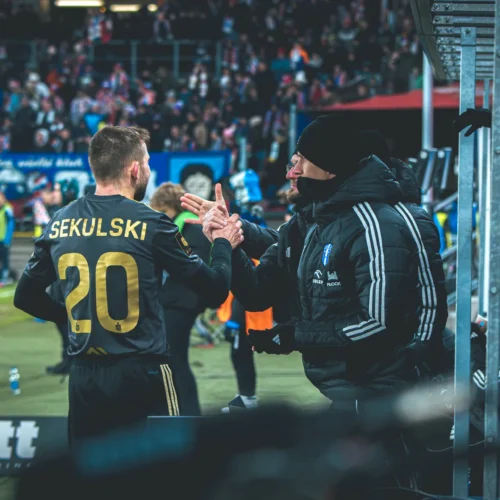 Na zdjęciu widać piłkarza z numerem 20, Sekulskiego, który wita się z trenerem podczas meczu, a w tle zebrani kibice.