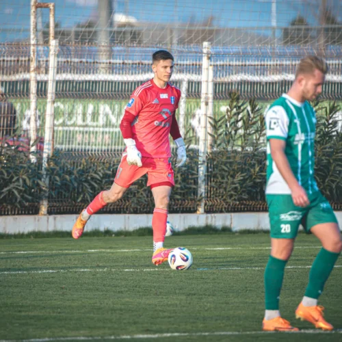 Na zdjęciu widać bramkarza w różowym stroju, który przygotowuje się do wykopnięcia piłki, podczas gdy jego kolega z drużyny w zielonej koszulce przechodzi w tle.