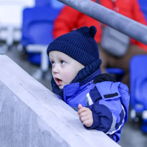Na zdjęciu widać małego chłopca w niebieskim płaszczu i czapce, który z zainteresowaniem obserwuje coś przed sobą, siedząc na trybunie stadionu.