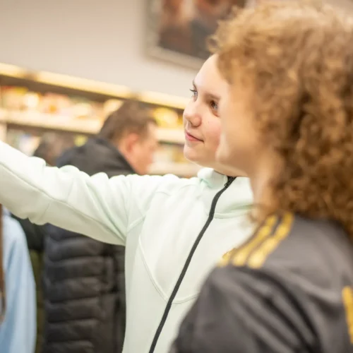 Dwie dziewczynki w sklepie robią selfie, podczas gdy w tle inni klienci przeglądają produkty.