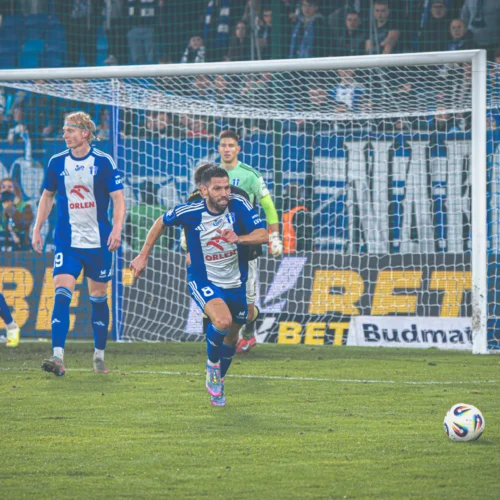 Na zdjęciu widać grupę piłkarzy w niebiesko-białych strojach, którzy świętują zdobycie bramki na boisku, z bramkarzem stojącym w tle.