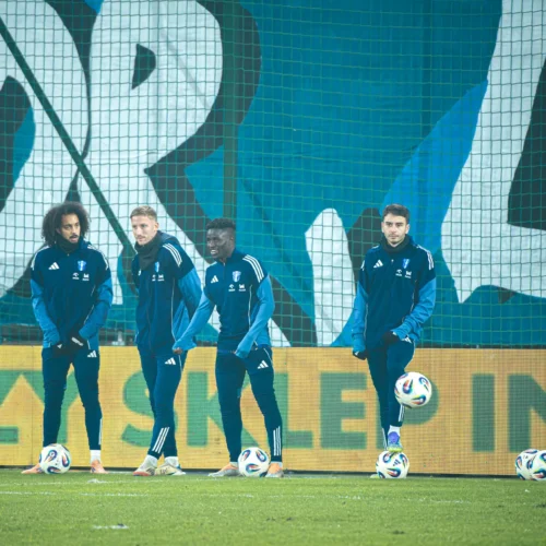 Na zdjęciu widać grupę piłkarzy w niebieskich dresach, którzy przygotowują się do treningu na boisku, w tle widoczna jest duża grafika z napisem.