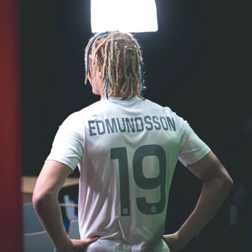 Na zdjęciu widoczna jest osoba w białej koszulce z napisem "EDMUNDSSON" i numerem "19", patrząca w stronę źródła światła.