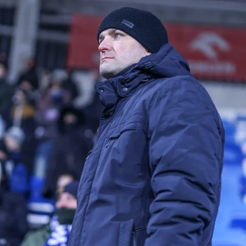 Mężczyzna w ciemnej kurtce i czapce obserwuje wydarzenie na stadionie, w tle widać zasiadających kibiców.