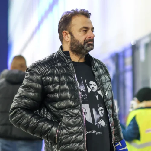 Mężczyzna w ciemnej kurtce i koszulce z nadrukiem stoi na tle oświetlonej ściany.