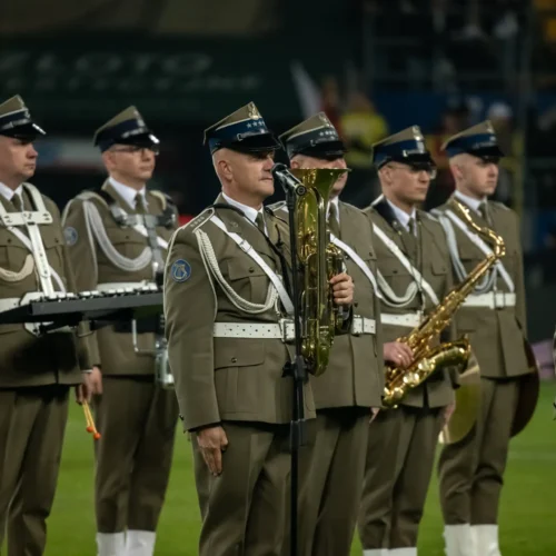 Grupa mężczyzn w mundurach wojskowych stoi w szeregu, trzymając instrumenty muzyczne podczas wydarzenia.