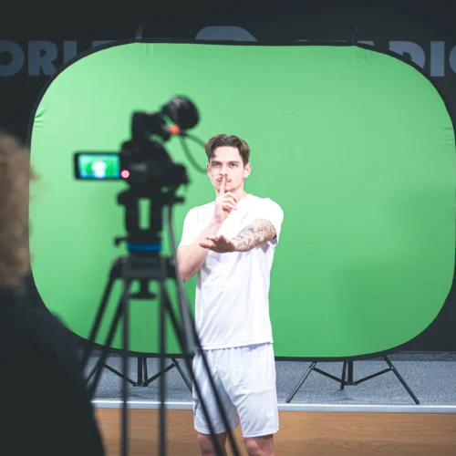 Na zdjęciu widoczny jest mężczyzna w białym stroju, który pozdrawia z gestem ciszy przed kamerą ustawioną na tle zielonego ekranu.