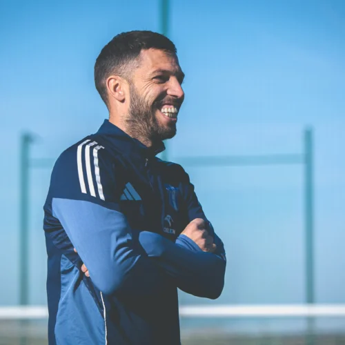 Każdy uśmiechnięty mężczyzna w sportowej odzieży stoi z założonymi rękami na tle boiska piłkarskiego.