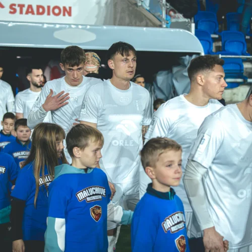 Na zdjęciu widać piłkarzy w białych strojach, którzy wychodzą na boisko, trzymając za ręce dzieci w niebieskich t-shirtach z napisem "Maluchowe Mistrzostwa".