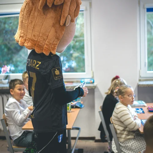 Na zdjęciu widoczna jest osoba w kostiumie z dużą, pluszową głową, która stoi w sali lekcyjnej, podczas gdy dzieci siedzą przy biurkach.