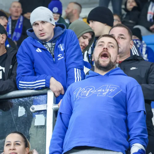 Na zdjęciu widać grupę kibiców, którzy intensywnie przeżywają wydarzenia na stadionie, z wyraźnym aplauzem i emocjami.