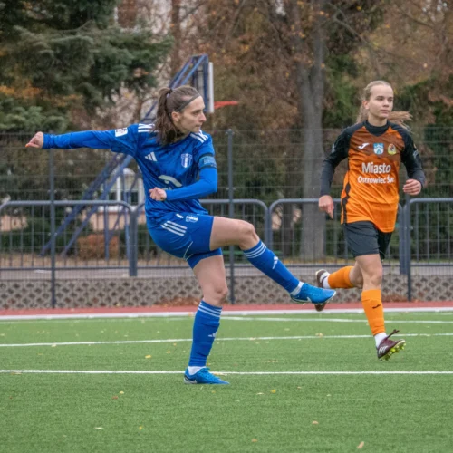 Na zdjęciu widać zawodniczkę w niebieskim stroju, która wykonuje kopnięcie piłki na boisku, podczas meczu piłki nożnej, a obok niej biegają inne zawodniczki.