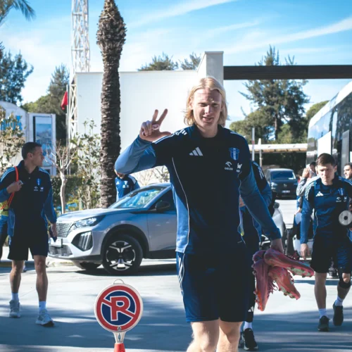 Na zdjęciu widać grupę sportowców wychodzących z autobusu, z jednym z nich pozującym z uśmiechem i gestem "victory".