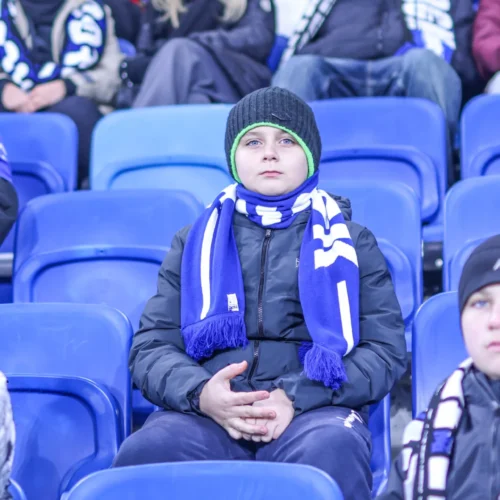 Na zdjęciu widać młodego kibica w szaliku siedzącego na trybunach, otoczonego innymi kibicami w zimowych ubraniach.