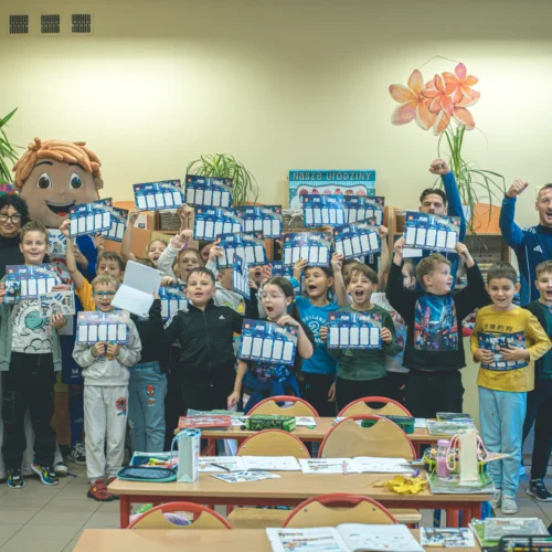 Na zdjęciu widać grupę dzieci w klasie trzymających kartki z planem, z uśmiechami na twarzach, obok nich stoją dorośli, w tym postać maskotki.