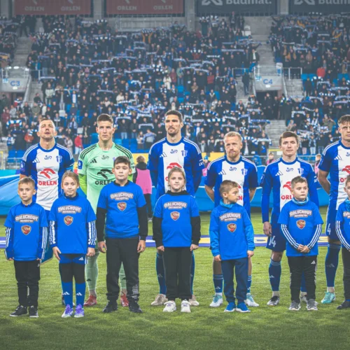 Na zdjęciu widoczny jest zespół piłkarski stojący na boisku w towarzystwie dzieci w strojach z logo klubu, przygotowujący się do rozpoczęcia meczu.