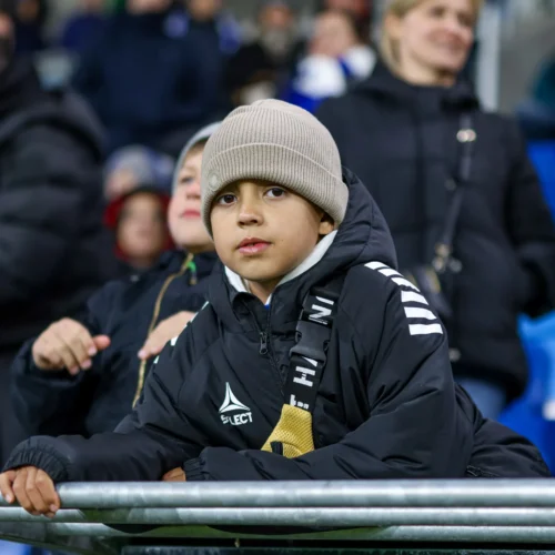 Chłopiec w zimowej kurtce i czapce siedzi na trybunach stadionu, skupiony na wydarzeniu sportowym.