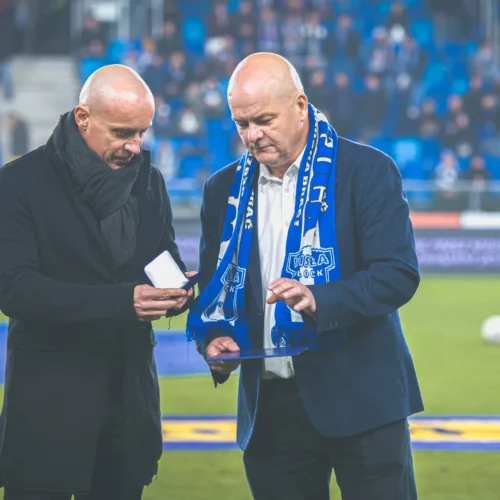 Na zdjęciu dwóch mężczyzn rozmawia na boisku sportowym, jeden trzyma małe pudełko, a drugi tablet, obaj są ubrani w eleganckie ubrania i szalik w barwach drużyny.