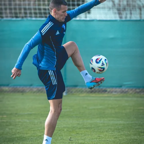 Na zdjęciu widać piłkarza w niebieskim stroju trenującego na boisku, wykonującego sztuczkę z piłką.