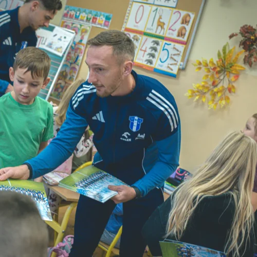 Na zdjęciu widać sportowca rozdającego dzieciom zdjęcia w klasie przedszkolnej.