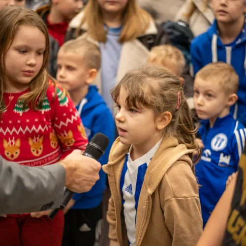 Dziewczynka w beżowej bluzie mówi do mikrofonu, otoczona przez grupę dzieci w różnych strojach sportowych i sweterkach.
