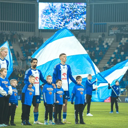 Na zdjęciu widoczni są piłkarze w niebiesko-białych strojach oraz dzieci w podobnych koszulkach, trzymający flagi, przed rozpoczęciem meczu na stadionie.