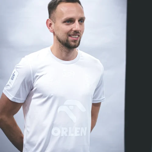 Zdjęcie przedstawia mężczyznę w białej koszulce z logo "ORLEN", uśmiechającego się podczas sesji zdjęciowej.