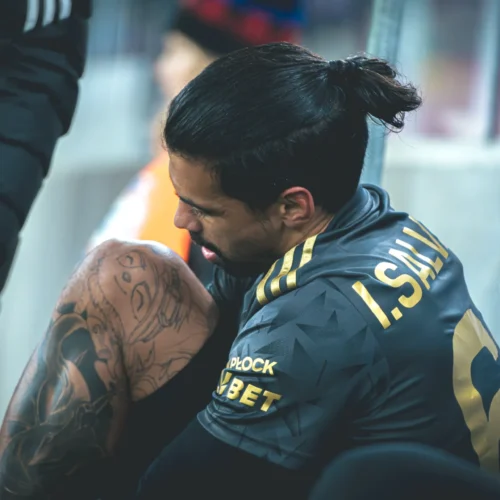 Na zdjęciu widoczny jest zawodnik znajdujący się na ławce rezerwowych, który z uwagą spogląda na swoją nogę z widoczną tatuażem.