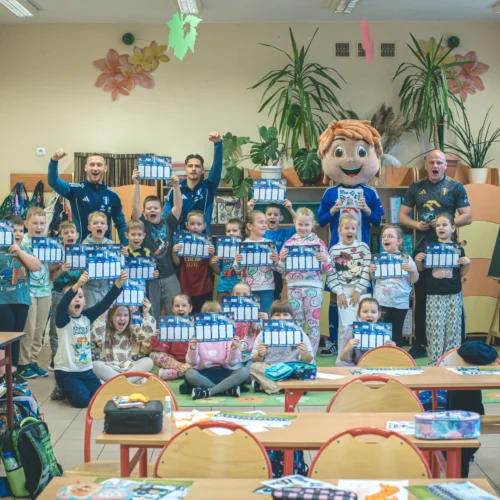 Na zdjęciu w szkolnej klasie grupa dzieci oraz dorośli trzymają wspólnie kalendarze, a w tle widać uśmiechnięte maskotki i radosną atmosferę.