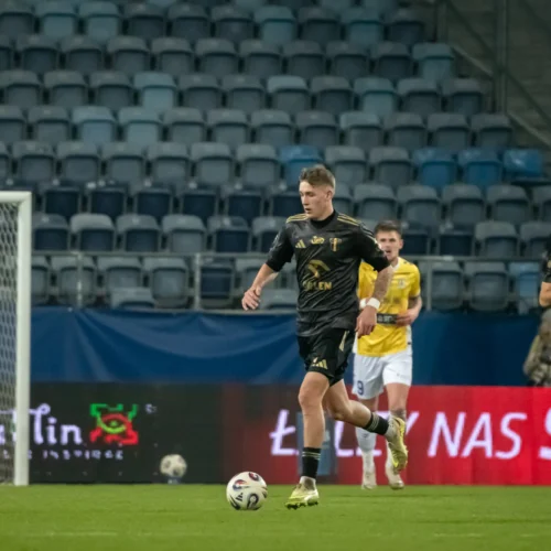 Na zdjęciu widać piłkarza w czarnym stroju biegnącego z piłką, podczas meczu piłkarskiego na pustym stadionie.