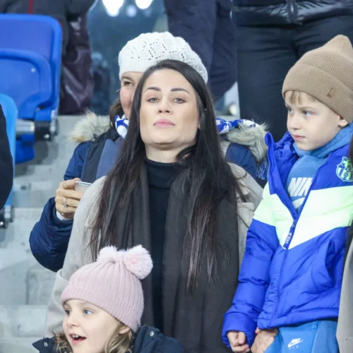 Na zdjęciu widać grupę osób na trybunach, w tym kobiety i dzieci, które wyglądają na zaangażowane w oglądanie wydarzenia sportowego.