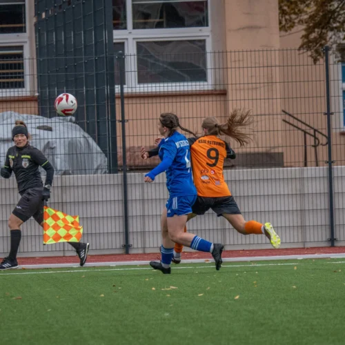 Na zdjęciu widoczna jest akcja podczas meczu piłki nożnej, w którym zawodniczki w niebieskich i pomarańczowych strojach biegają za piłką, a sędzia pomocniczy z flagą obserwuje sytuację.