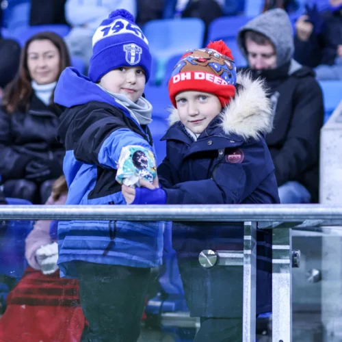 Na zdjęciu widać dwóch małych chłopców w ciepłych ubraniach i czapkach, którzy uśmiechają się, trzymając się za ręce na tle tłumu na trybunach stadionu.