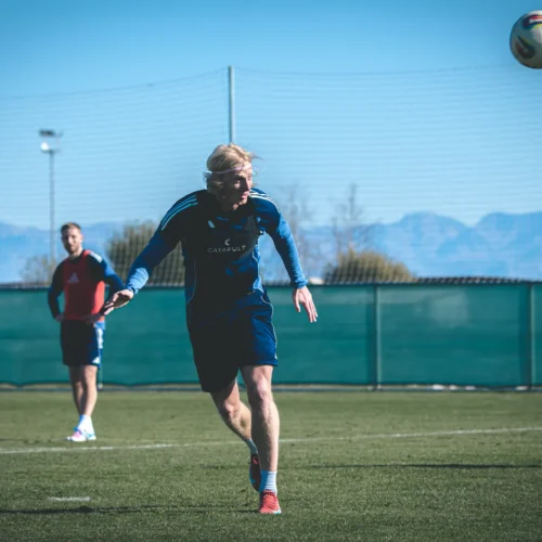 Na zdjęciu widać zawodników trenujących na boisku piłkarskim, z jednym z nich dynamicznie podążającym za piłką.