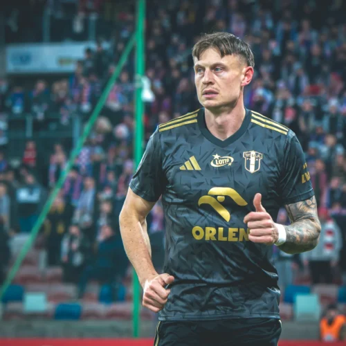 Na zdjęciu widać piłkarza w czarnym stroju z logo drużyny, który stoi na boisku z podniesioną ręką, w tle widać tłum kibiców.