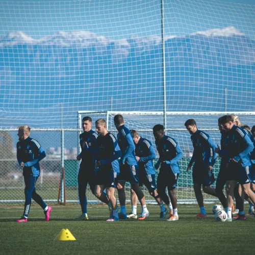 Zespół piłkarski w niebieskich strojach trenuje na boisku, z górskim krajobrazem w tle.